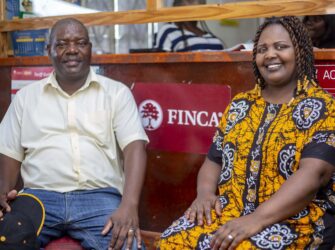 Man wearing white shirt and woman wearing yellow printed dress sitting on a bench in front of a FINCA Sign. both are smiling.