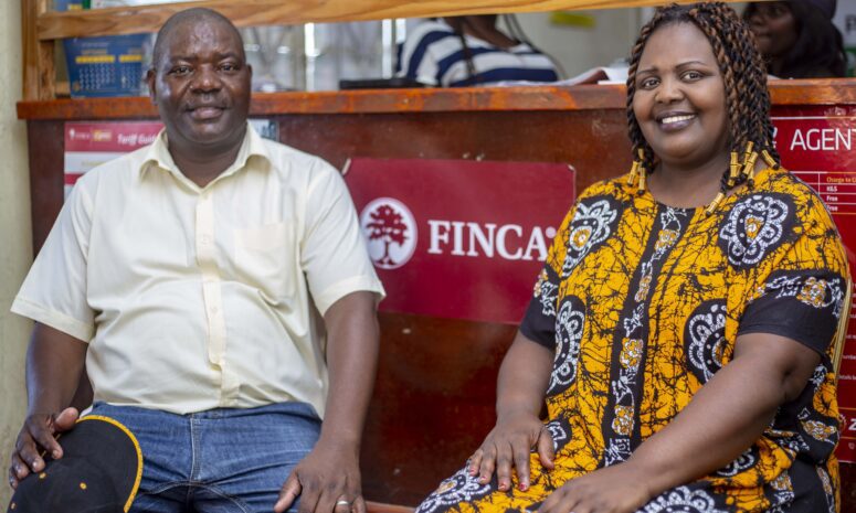 Man wearing white shirt and woman wearing yellow printed dress sitting on a bench in front of a FINCA Sign. both are smiling.