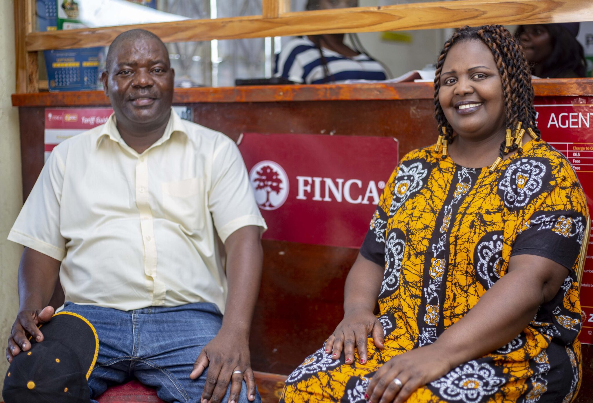 Man wearing white shirt and woman wearing yellow printed dress sitting on a bench in front of a FINCA Sign. both are smiling.