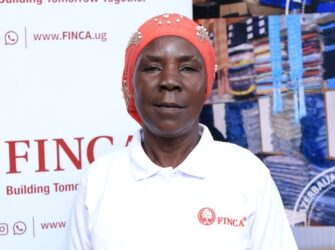 FINCA Uganda Brand Ambassador Janet Mudoola standing in front of a FINCA banner. She is wearing a red head covering and a FINCA branded collared shirt.