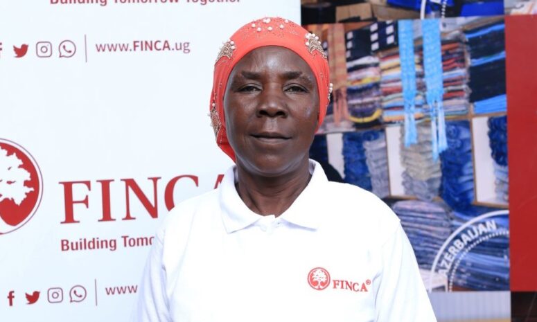 FINCA Uganda Brand Ambassador Janet Mudoola standing in front of a FINCA banner. She is wearing a red head covering and a FINCA branded collared shirt.