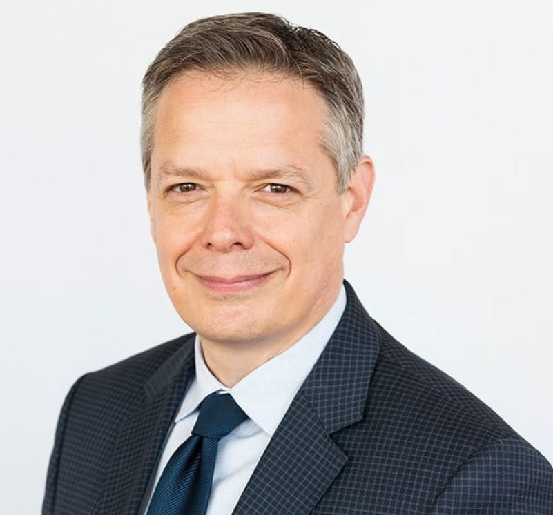Head and shoulders image of Dan Smith, smiling, wearing a dark suit and tie with a light colored shirt.