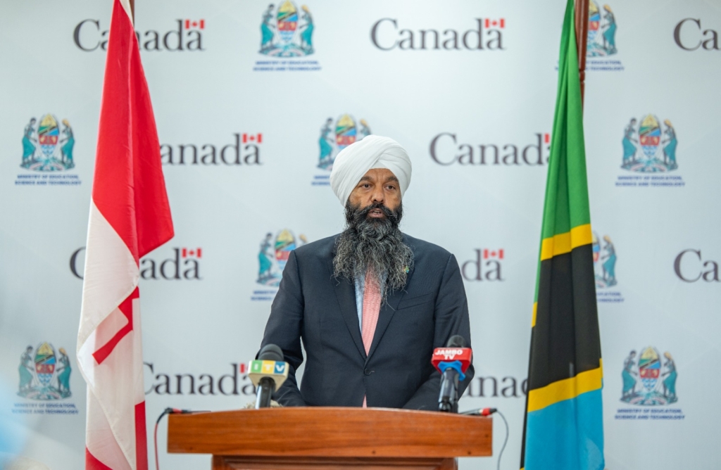 The honorable Randeep Sarai, Secretary of State for International Development, Government of Canada, speaks in Tanzania. He is wearing a dark suit, light blue shirt and pink tie and is standing at a podium. There is a Canadian flag on the left of the podium and a Tanzania flag on the right. Behind him is a white backdrop imprinted with the word Canada. 