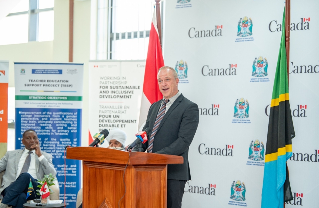 FINCA Canada Executive Director Drew Boshell speaking in Tanzania. He is standing at a podium with a Canada flag to the left and a Tanzania flag to the right. A white backdrop with the word Canada imprinted on it is behind him. He is wearing a dark suit, light shirt and striped tie. 